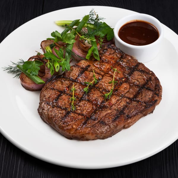 Grilled steak with herbs, roasted onions, and a side of dark sauce on a white plate.