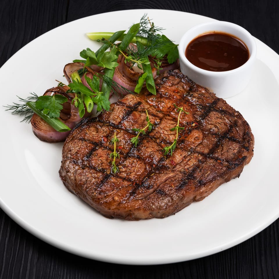 Grilled steak with herbs, roasted onions, and a side of dark sauce on a white plate.