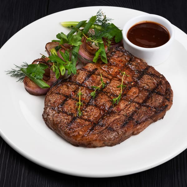 Grilled steak with herbs, roasted onions, and a side of dark sauce on a white plate.
