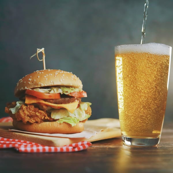 A crispy chicken burger with cheese, lettuce, and tomato next to a glass of beer being poured.